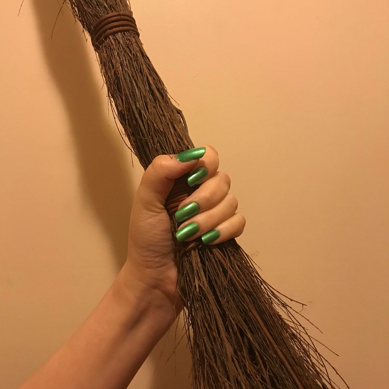 May include: A person holding a brown broom with a hand painted with green nail polish.