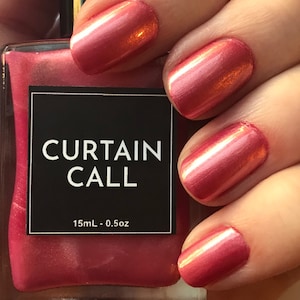 May include: A bottle of nail polish with the text "CURTAIN CALL" on a black label. The nail polish is a shimmery, reddish-pink color. The nails on the hand are painted with the same color.