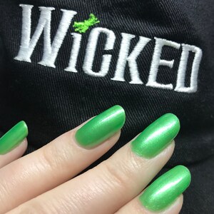 May include: A hand with green nail polish, the nails are shiny and have a metallic finish. The hand is resting on a black fabric with the word "WICKED" embroidered in white.