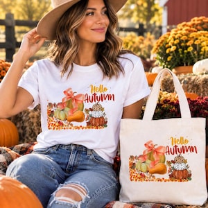 Puede incluir: Una camiseta blanca y una bolsa de tela a juego, ambas con un estampado gráfico de calabazas coloridas, hojas de otoño y el texto Hello Autumn en letra naranja y marrón. El diseño incluye una pila de calabazas y un lazo decorativo.