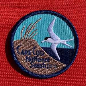 May include: Embroidered patch featuring a coastal scene with a flying bird, dune grass, and the text "CAPE COD National Seashore." The patch has a blue, teal, and tan color scheme, with a dark blue border. The background is red.