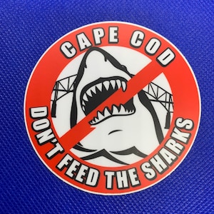 May include: A red circle with a white outline and the text "CAPE COD DON'T FEED THE SHARKS" around the edge. A black and white illustration of a shark with its mouth open is in the center of the circle. A red line is drawn across the shark's mouth.