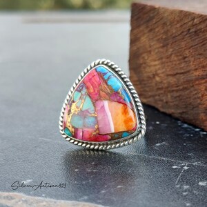 May include: A triangular silver ring with a colorful inlay of turquoise, pink, orange, and gold. The ring has a rope-like design around the edge.