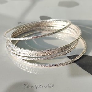 May include: A stack of silver bangles with a hammered texture. The bangles are thin and round, reflecting light. The image is taken on a light-colored surface. The text "Silver Artisan 925" is in the bottom right corner.