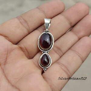 May include: A silver pendant featuring two dark red gemstones. The top stone is oval, and the bottom is teardrop-shaped. The pendant has a loop for a chain. The text "SilverArtisan925" is visible on the bottom right.