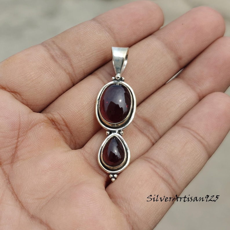 May include: A silver pendant featuring two dark red gemstones. The top stone is oval, and the bottom is teardrop-shaped. The pendant has a loop for a chain. The text "SilverArtisan925" is visible on the bottom right.