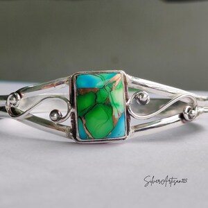May include: A silver cuff bracelet with a rectangular turquoise and green stone center. The bracelet has a decorative, openwork design with swirling details and a polished finish. The stone has copper-colored inclusions.