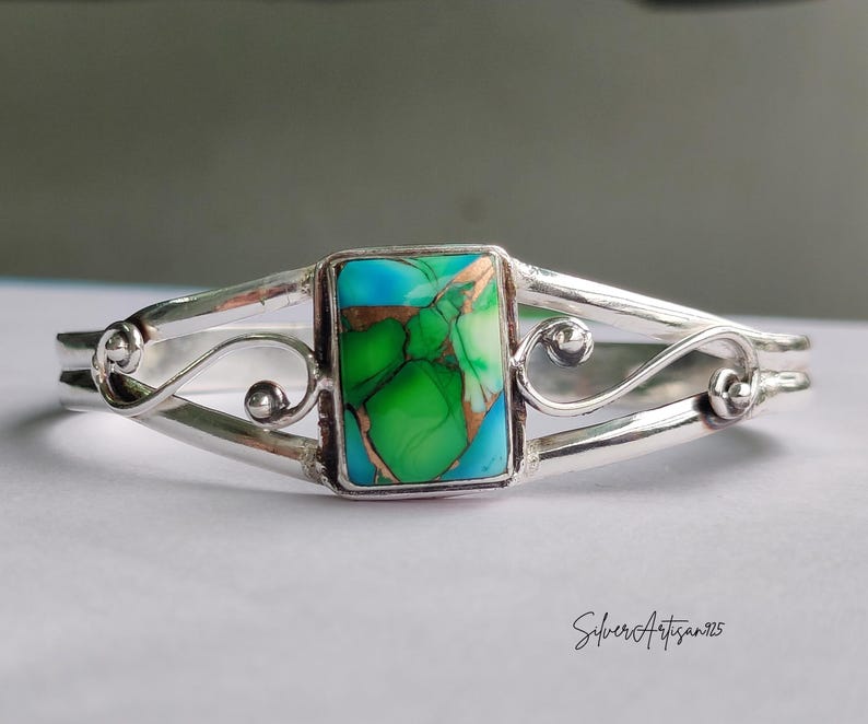 May include: A silver cuff bracelet with a rectangular turquoise and green stone center. The bracelet has a decorative, openwork design with swirling details and a polished finish. The stone has copper-colored inclusions.