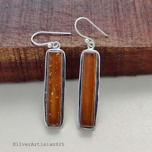 May include: A pair of rectangular earrings with a silver frame. The earrings feature a warm amber-colored center with gold flecks. The earrings are displayed on a wooden surface.