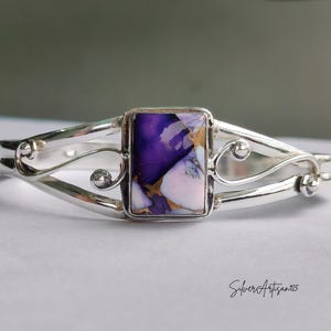 May include: A silver cuff bracelet with a rectangular, multi-colored stone. The stone features shades of purple, white, and gold. The bracelet has a decorative, openwork design with curved lines and small, round accents. The text "SilverArtisan925" is visible.