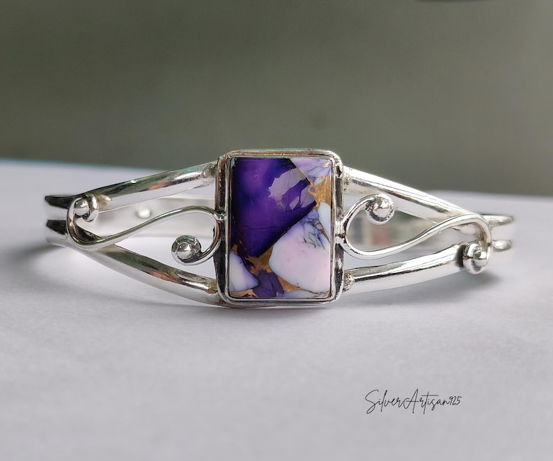 May include: A silver cuff bracelet with a rectangular, multi-colored stone. The stone features shades of purple, white, and gold. The bracelet has a decorative, openwork design with curved lines and small, round accents. The text "SilverArtisan925" is visible.