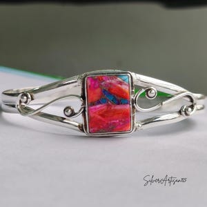 May include: A silver bracelet with a rectangular, multi-colored stone at the center. The stone features shades of red, pink, and blue. The bracelet has a decorative, swirling design on each side of the stone. The text "Silver Artisan925" is visible.