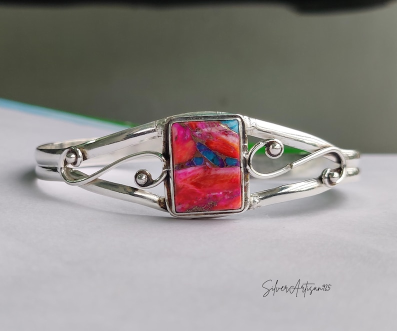 May include: A silver bracelet with a rectangular, multi-colored stone at the center. The stone features shades of red, pink, and blue. The bracelet has a decorative, swirling design on each side of the stone. The text "Silver Artisan925" is visible.