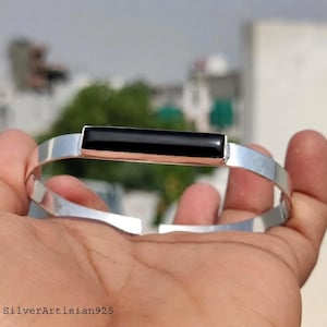 May include: A silver bracelet with a rectangular black onyx stone set in the center. The bracelet has a simple, modern design and is held in a hand. The text "SilverArtisian925" is at the bottom.