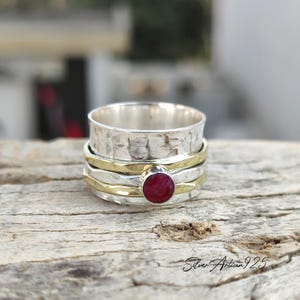 May include: A silver spinner ring with a hammered texture and gold bands. A round, red gemstone is set in the center. The ring is displayed on a wooden surface. The text "Silver Artisan 925" is visible.