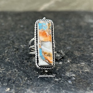 May include: A silver ring with a rectangular turquoise, orange, and white stone set in a silver bezel. The ring has a rope-like design around the edge of the bezel.