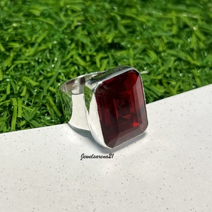 May include: A silver ring with a large, rectangular, deep red gemstone. The ring has a wide band and a simple, modern design. The gemstone is faceted, catching the light and creating a sparkling effect. The ring is displayed on a white surface with green grass in the background.