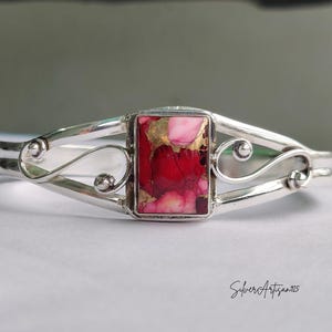 May include: A silver cuff bracelet with a rectangular, red and pink stone center. The stone has gold flecks. The bracelet has a decorative, swirling design on each side of the stone.