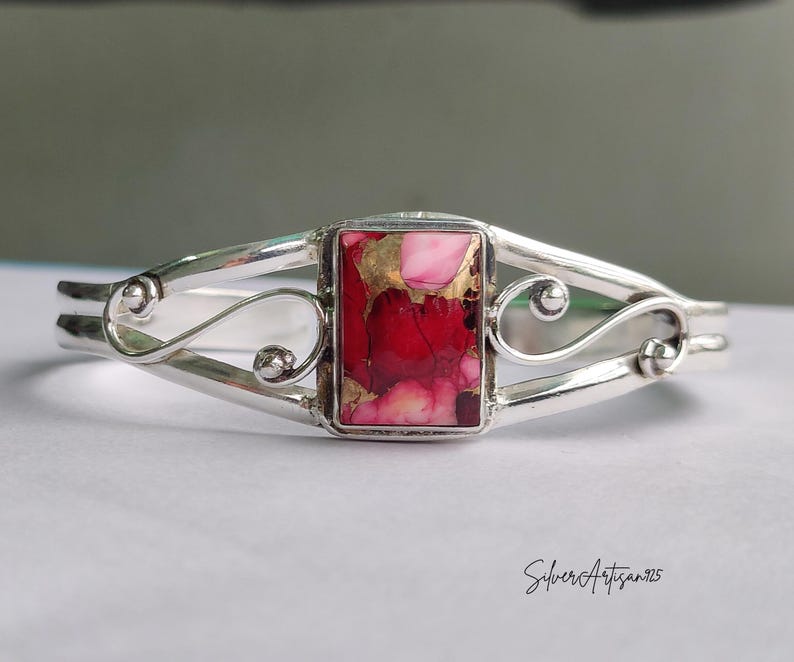 May include: A silver cuff bracelet with a rectangular, red and pink stone center. The stone has gold flecks. The bracelet has a decorative, swirling design on each side of the stone.