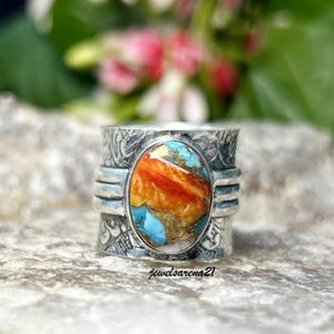 May include: A silver ring with a large oval-shaped turquoise stone set in the center. The stone has a mix of orange, yellow, and blue colors. The ring band is textured with a floral design.