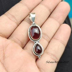 May include: A silver pendant featuring two red gemstones, an oval and a teardrop shape. The pendant is displayed against a hand. The gemstones are set in silver.
