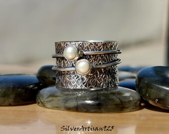 Könnte beinhalten: Ein silberner Spinnerring mit zwei weißen Perlen. Der Ring hat ein strukturiertes Design und ist aus Sterlingsilber.