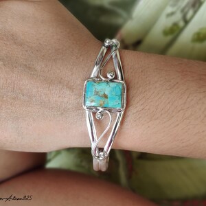 May include: A silver cuff bracelet with a rectangular turquoise stone at the center. The bracelet has an open design with decorative swirls and small silver balls. The turquoise stone has a mottled blue-green appearance.