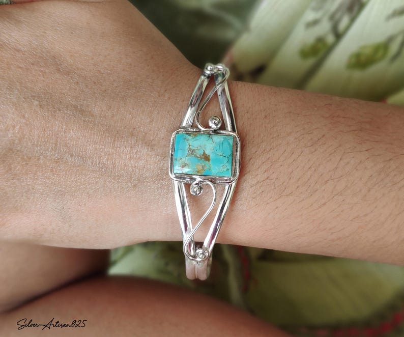 May include: A silver cuff bracelet with a rectangular turquoise stone at the center. The bracelet has an open design with decorative swirls and small silver balls. The turquoise stone has a mottled blue-green appearance.