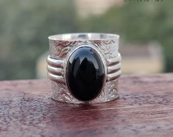 Puede incluir: Un anillo de plata con una piedra ovalada de ónix negro. El anillo tiene una banda ancha con un diseño grabado intrincado.