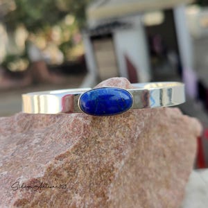 May include: A silver cuff bracelet with a polished finish. The bracelet features a large, oval-shaped, deep blue gemstone set in the center. The bracelet is resting on a rough, reddish-brown stone surface.