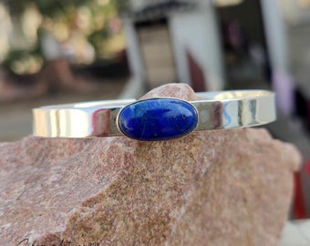 May include: A silver cuff bracelet with a polished finish. The bracelet features a large, oval-shaped, deep blue gemstone set in the center. The bracelet is resting on a rough, reddish-brown stone surface.