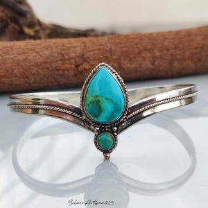 May include: A silver cuff bracelet featuring a V-shaped design with a central pear-shaped turquoise stone and a smaller round turquoise stone below it. The silver band has a twisted rope texture along the edges and a polished finish throughout.