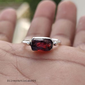 May include: A silver ring featuring a rectangular, deep red gemstone. The ring has a simple band with small, round accents on either side of the stone. The gemstone has a rough, natural texture. The text "SilverArtisian925" is at the bottom.