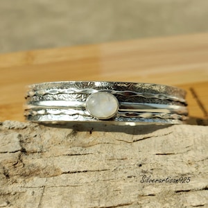 May include: A silver-toned bracelet with an oval, light-colored stone at the center. The bracelet has a textured, layered design with etched patterns. The text "Silverartisan925" is visible on the wooden surface.