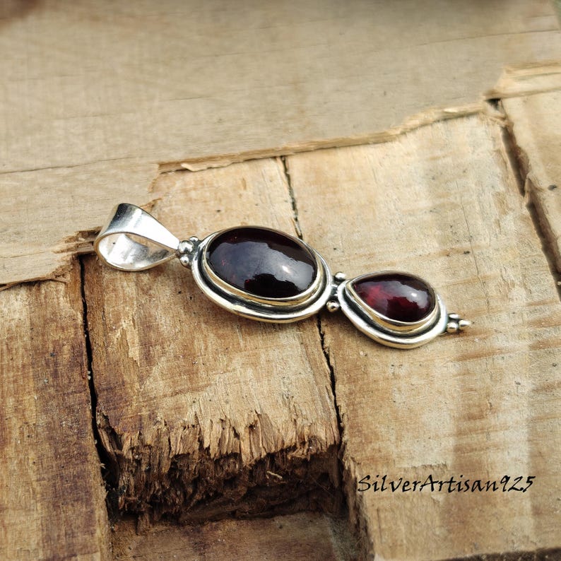May include: A silver pendant with two red garnet gemstones, one oval and one teardrop-shaped. The gemstones are set in a silver bezel. The pendant is displayed on a wooden surface.
