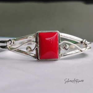 May include: A silver bracelet with a rectangular, bright red stone at the center. The bracelet has a decorative, open design with curved lines and small, round accents. The text "SilverArtisan925" is written in cursive on the bottom right.