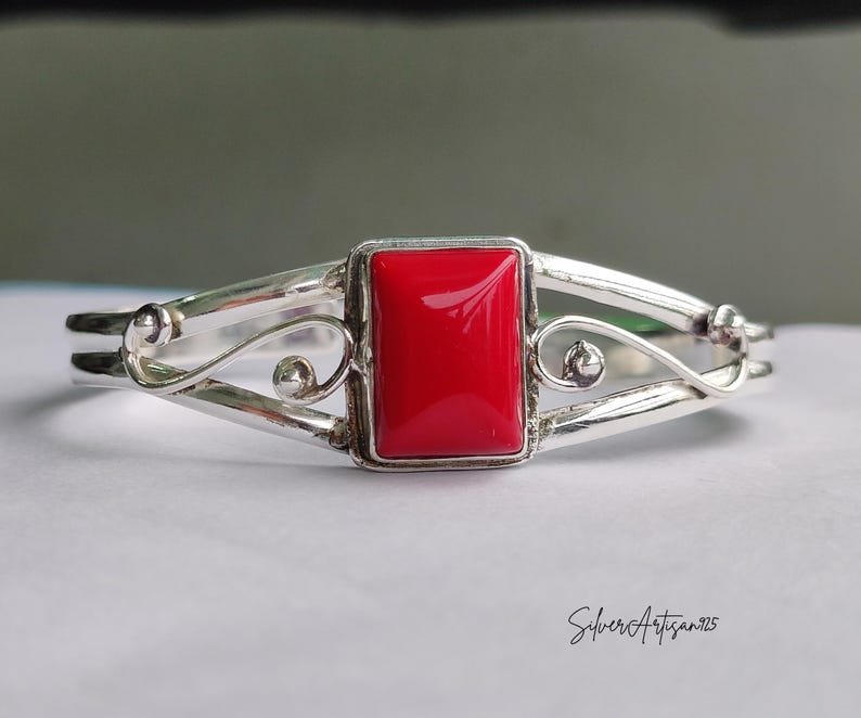 May include: A silver bracelet with a rectangular, bright red stone at the center. The bracelet has a decorative, open design with curved lines and small, round accents. The text "SilverArtisan925" is written in cursive on the bottom right.