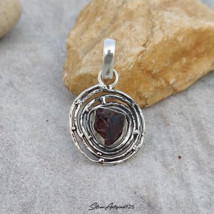 May include: A silver pendant featuring a raw, dark red garnet stone set within a spiral design. The pendant has a loop for a chain and is displayed on a neutral surface. The text "Silver Artisan925" is at the bottom.