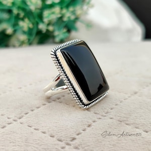 May include: A silver ring with a black rectangular onyx stone set in a rope-like border.