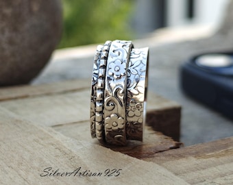May include: A silver spinning ring with a floral design. The ring is made of three bands, each with a different floral pattern. The ring is on a wooden surface.