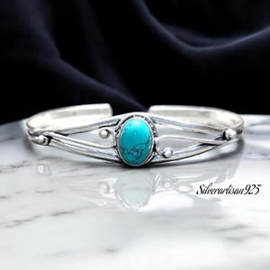 May include: A silver bangle bracelet featuring a turquoise stone set in the centre. The bracelet has a delicate, intricate design with a curved shape.