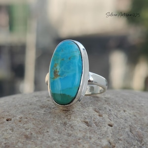 May include: A silver ring with a large oval turquoise stone set in a bezel setting. The stone has a blue and green color with brown veins.