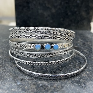 May include: A set of six silver bangles with intricate floral and wave designs. Three of the bangles feature a light blue opal stone.