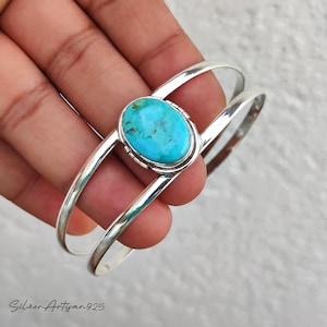 May include: A silver cuff bracelet featuring a large, oval turquoise stone. The bright blue stone is set in a silver bezel and the bracelet has a split design. The bracelet is shown on a hand.