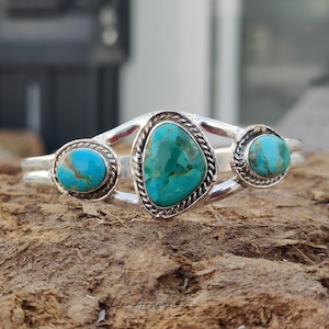 May include: A silver cuff bracelet featuring three turquoise stones with brown matrix patterns. The central stone is pear-shaped, flanked by two smaller round stones, all set in twisted silver wire frames. The text Silver Artisan925 is visible.