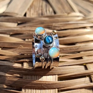 Peut inclure: Une bague rotative en argent ornée de deux opales rondes irisées et d'une pierre centrale ronde en labradorite bleue, agrémentée de petites perles en métal doré. L'anneau a une finition polie et porte l'inscription Jewelsarena21.