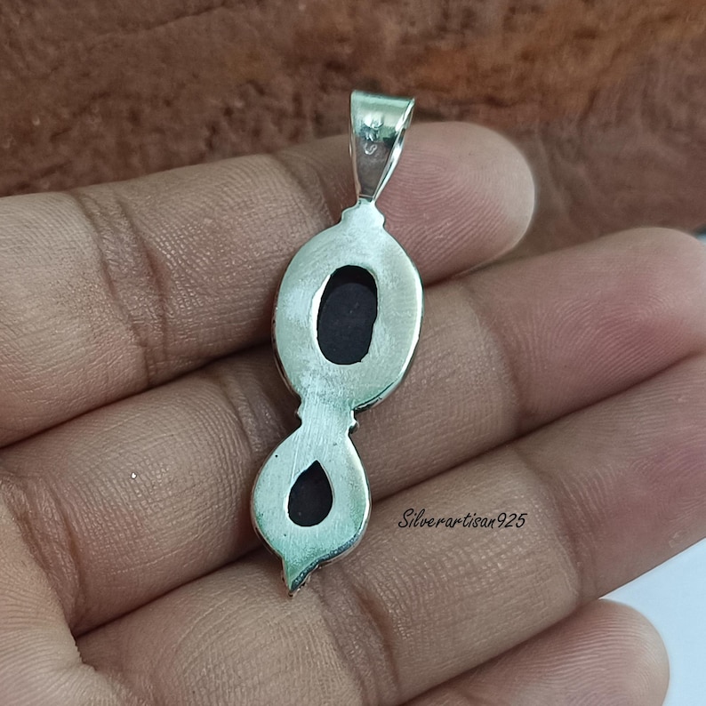 May include: A silver pendant with an elongated, figure-eight design. The pendant features two oval-shaped, dark-coloured stones set in a silver frame. The text "Silverartisan925" is visible on the right side of the pendant.