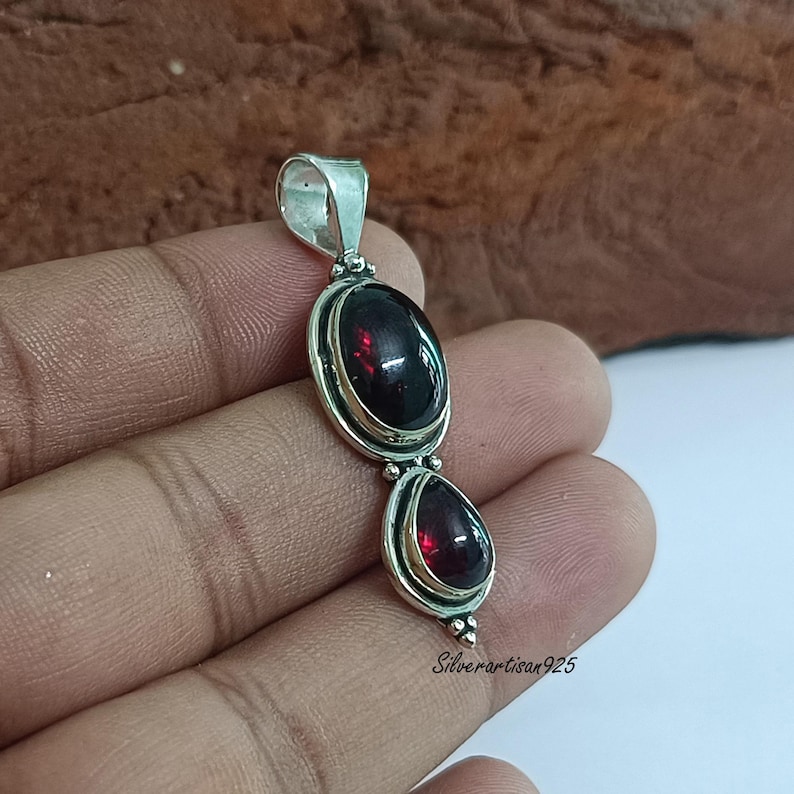 May include: A silver pendant with two dark red gemstones, one oval and one teardrop-shaped. The gemstones are set in a silver frame. The pendant has a loop for a chain. The text "Silverartisan925" is visible on the pendant.