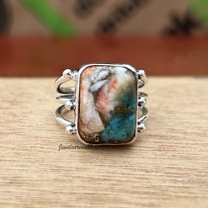 May include: A silver ring with a rectangular turquoise, pink, and white stone set in a bezel setting. The ring has a triple band design with small beads on the sides.