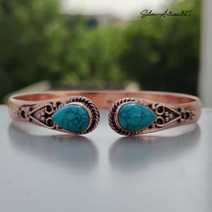 May include: Copper cuff bracelet with turquoise teardrop-shaped stones. The bracelet features intricate decorative details and the text "Silver Artisan925". The open design allows for easy wear.